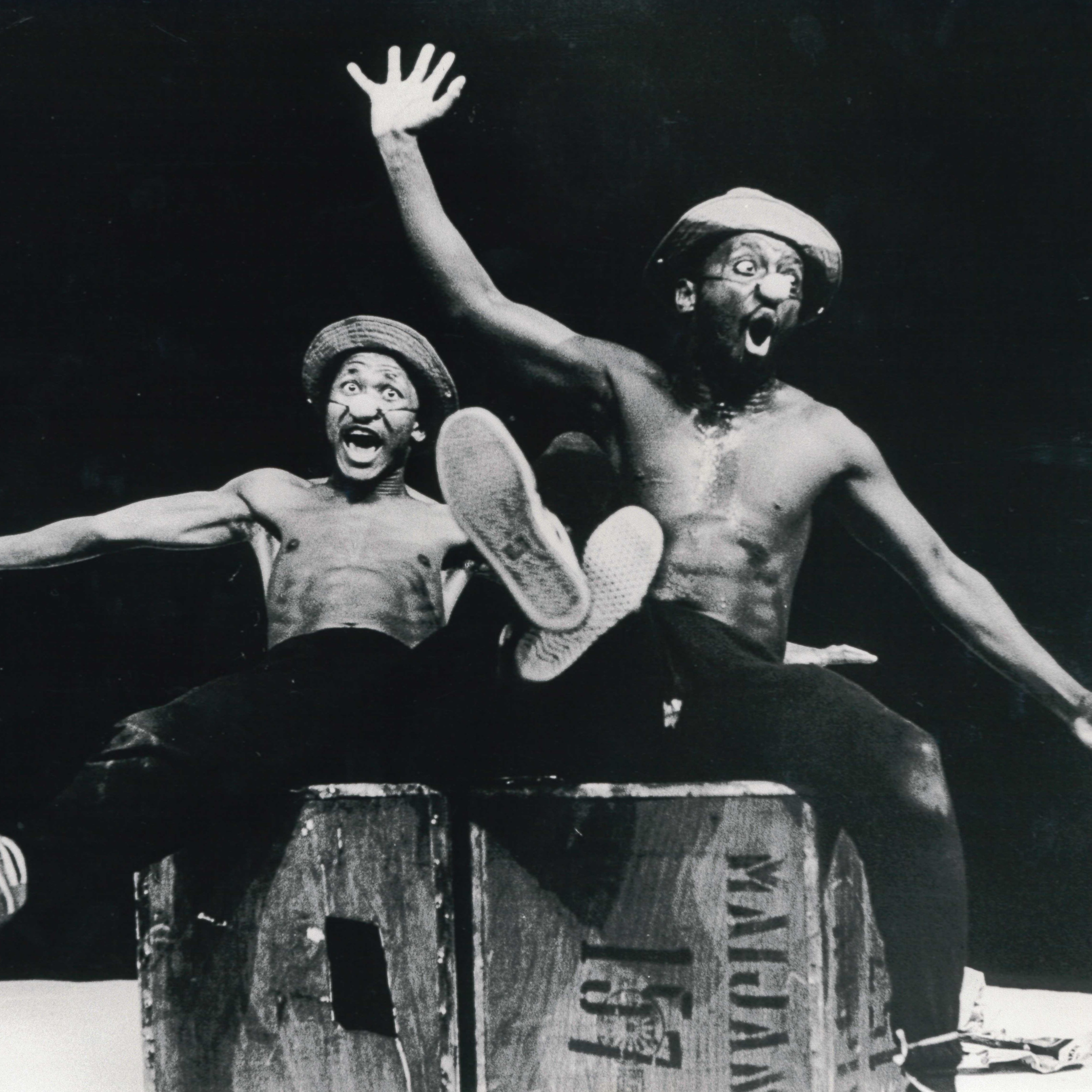 Black and white archival image of Percy Mtwa and Mbongeni June in Woza Albert On Stage 1985. Shows two actors sitting on a box together laughing with legs and arms outstretched.