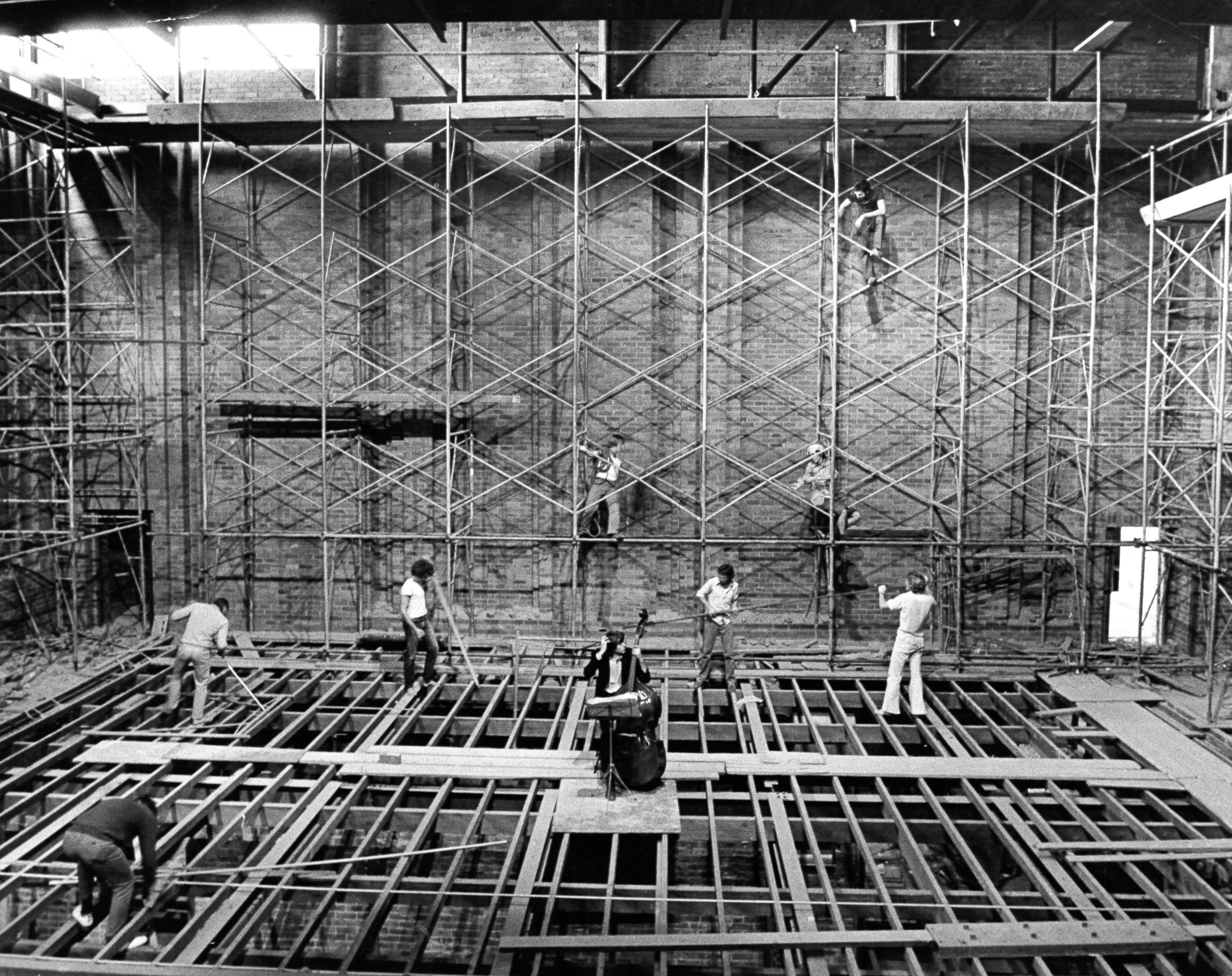 Black and white archival image of the Everest Theatre construction with a cellist surrounded by 7 construction workers.