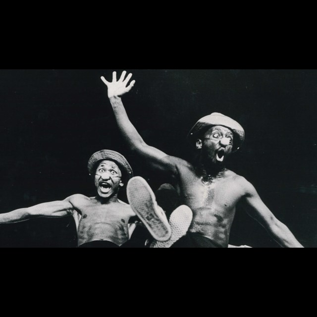 Black and white archival image of Percy Mtwa and Mbongeni June in Woza Albert On Stage 1985. Shows two actors sitting on a box together laughing with legs and arms outstretched.