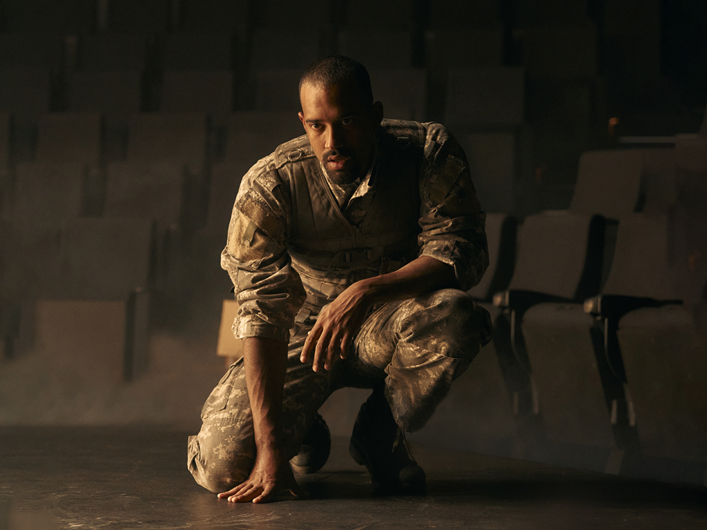 Promotional image of Othello Symposium for Seymour Centres Arts Education Program depicting a man in camouflage army uniform crouching and looking past the camera.
