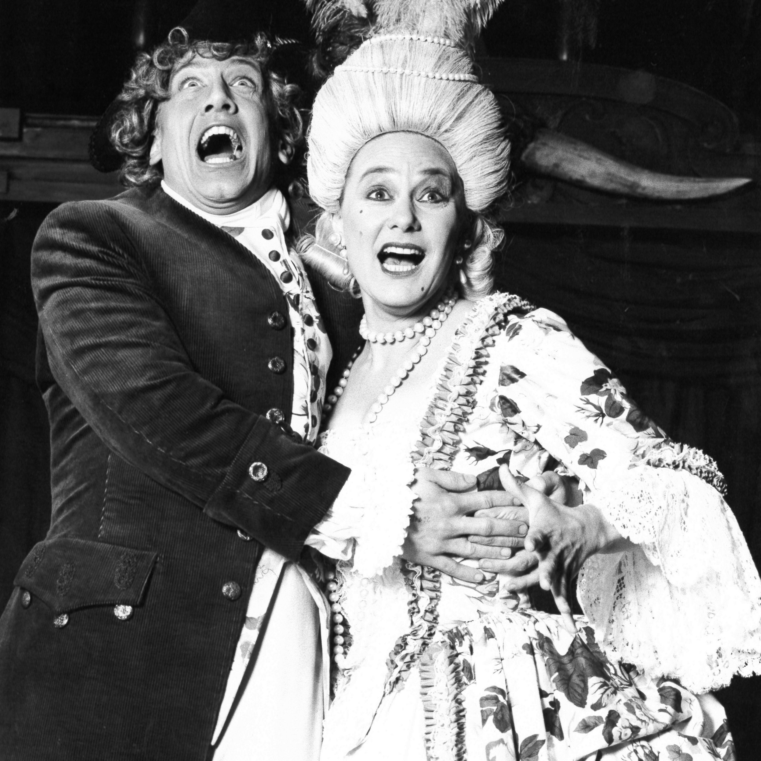 Black and white archival image of two actors hugging and signing out to the audience.