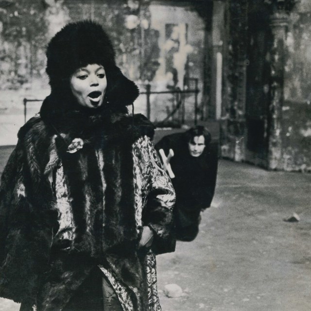 Black and white archival image of two actors, the female actor in the foreground singing and the male actor in the background creeping up behind.