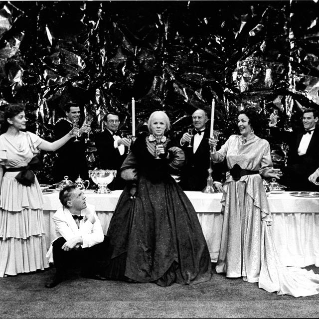 Black and white archival image depicting a dinner table set with 8 actors in ball gowns and tuxedoes all raising a toast with their drinking glasses.