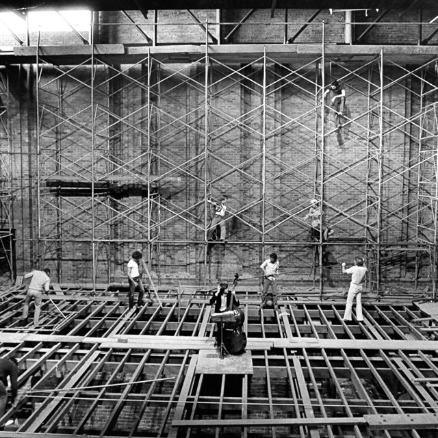 Black and white archival image of the Everest Theatre construction with a cellist surrounded by 7 construction workers.