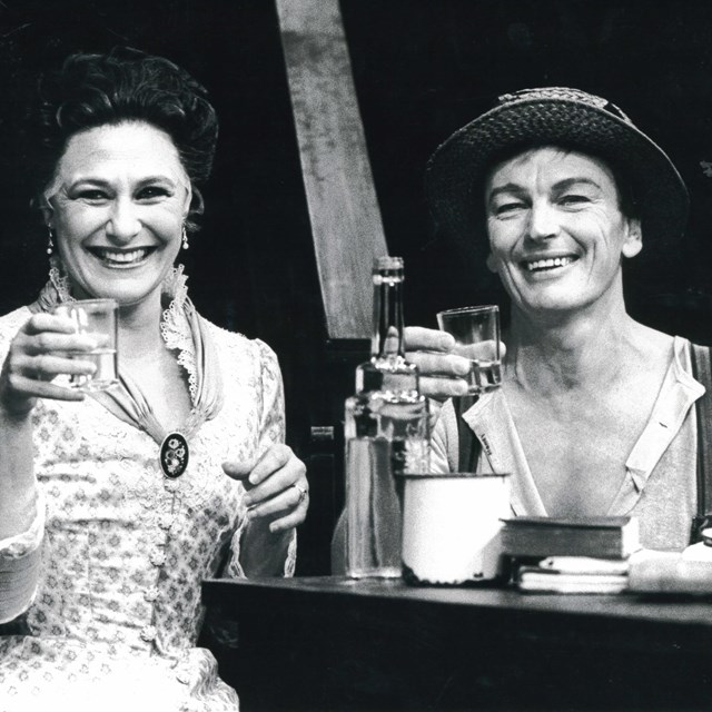 Black and white archival image of John Bell and Deidre Rubenstein in Wild Honey from 1986. Image shows both actors sitting at a table, smiling at the camera holding drinks in a sort of cheers with teh audience.